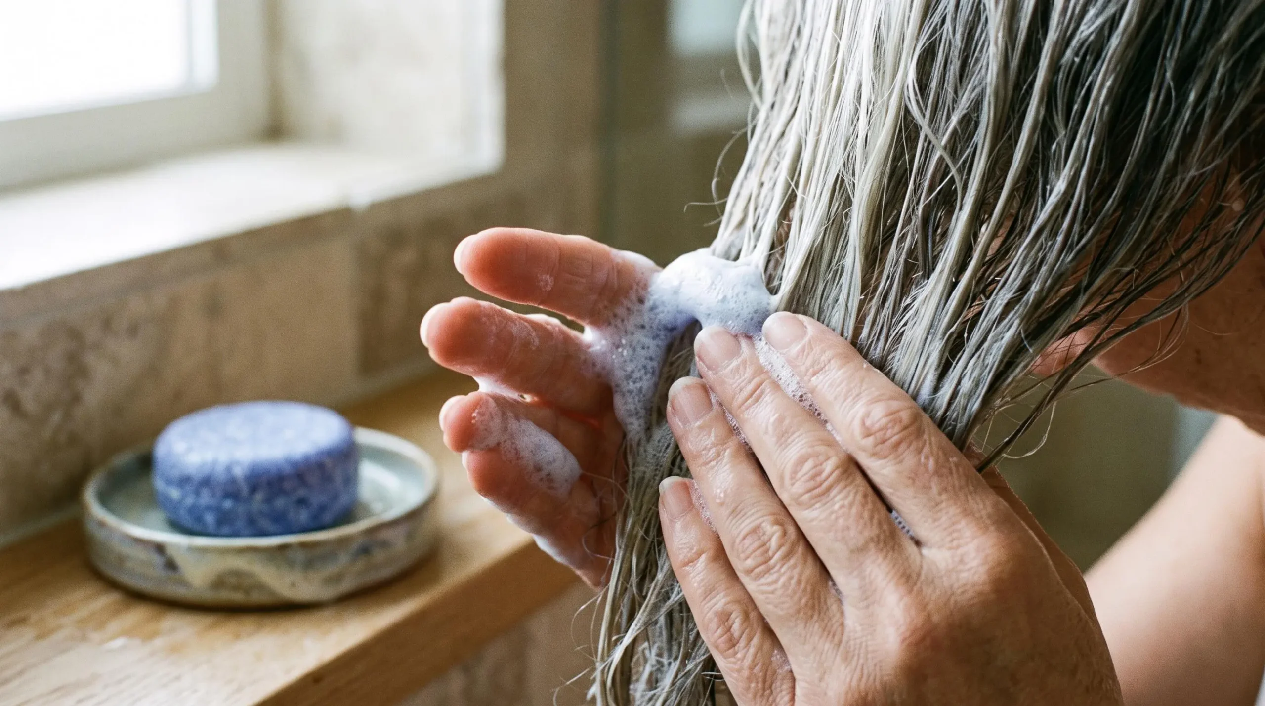 Mains appliquant de la mousse de shampoing sur les longueurs de cheveux blancs, le galet bleu posé en arrière-plan